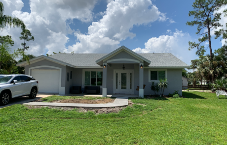 Completed Single-Story Home with Front Porch and Lawn A finished single-story home with a light gray exterior and a simple front porch. The house has a two-car driveway with an SUV parked on the left side. There is a well-maintained green lawn in the foreground and palm trees in the background. The sky is bright with scattered clouds.